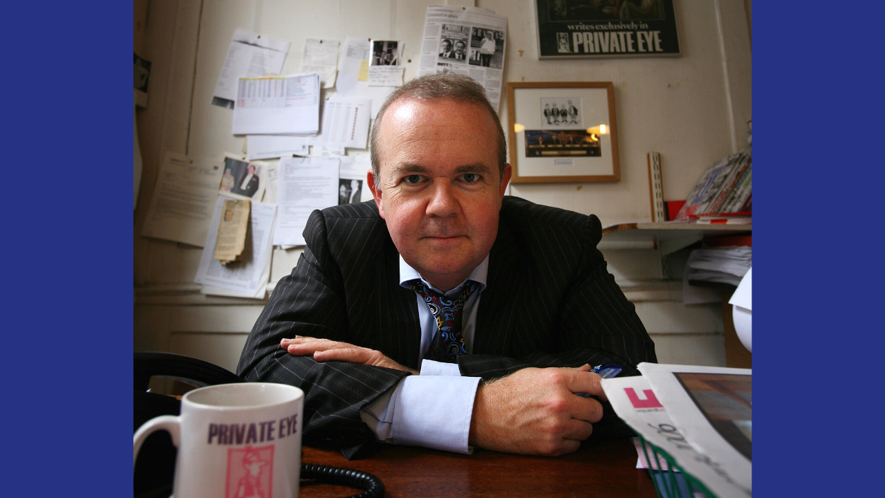 A photo of a white man with short light coloured hair leaning on a desk. He wears a pinstripe suit. In the foreground there is a stack of newspapers and a mug with red text reading 'Private Eye'. Papers are tacked to the wall in the background. The photo is framed with a blod blue surround.